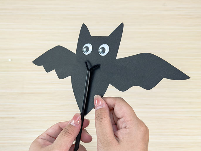 Two hands hold a simple bat puppet against a light wood-grain background. The bat is made of black construction paper with large googly eyes and a piece of black yarn attached below its face. A black stick or straw is visible behind the yarn, suggesting it's used as a handle for the puppet.