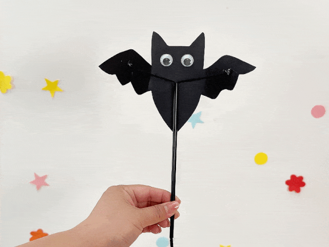 A hand holds a completed bat puppet against a white background decorated with small, colorful felt shapes of stars, circles, and flowers. The bat is made of black construction paper, has large googly eyes, and is attached to a black stick or straw handle. Black yarn connects the handle to the bat's wings, allowing them to be manipulated.