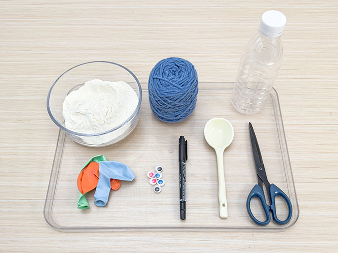 An assortment of craft supplies is arranged on a clear rectangular tray placed on a light wood-grain surface. The tray contains a clear bowl of flour, a ball of blue yarn, an empty clear plastic bottle, a white plastic spoon, a black marker, a pair of blue-handled scissors, a few small deflated balloons in various colors, and a pile of small googly eyes. These items appear to be gathered for a craft project.