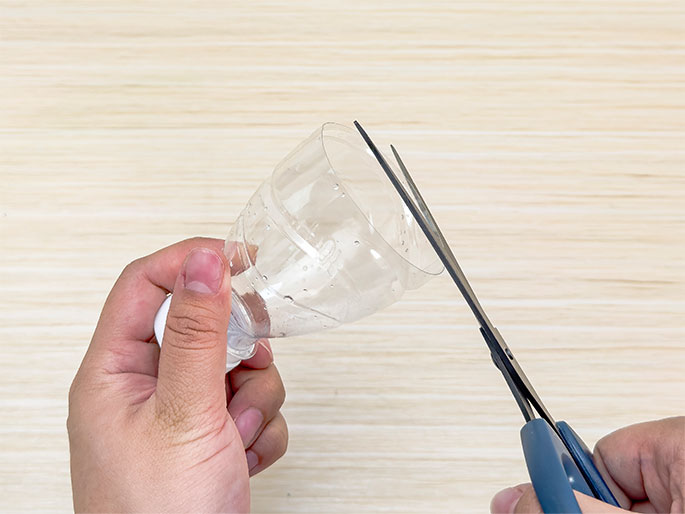 Two hands are shown cutting a clear plastic bottle against a light wood-grain background. One hand holds the bottle upside down near its cap, while the other hand uses a pair of blue-handled scissors to cut off the top portion of the bottle.