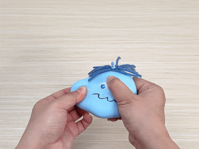 Two hands hold and gently manipulate a small, light blue, balloon-like figure against a light wooden background. The figure has a single googly eye, a wavy line for a mouth, and a tuft of blue yarn tied at the top to resemble hair. The hands press and shape the figure, demonstrating its soft and pliable nature.