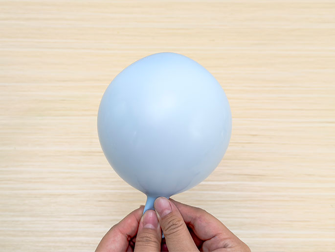 Two hands pinch the neck of a pale blue, uninflated balloon against a light wood-grain background. The balloon is round and smooth, and the hands are positioned near the balloon's opening, presumably preparing to inflate it.