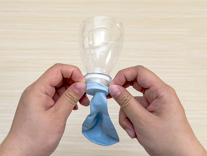 Against a light wood-grain background, two hands are shown attaching a deflated pale blue balloon to the open top of a clear plastic bottle. The top of the bottle appears to have been cut off, and the hands are stretching the balloon's opening over the bottle's rim to create a seal.