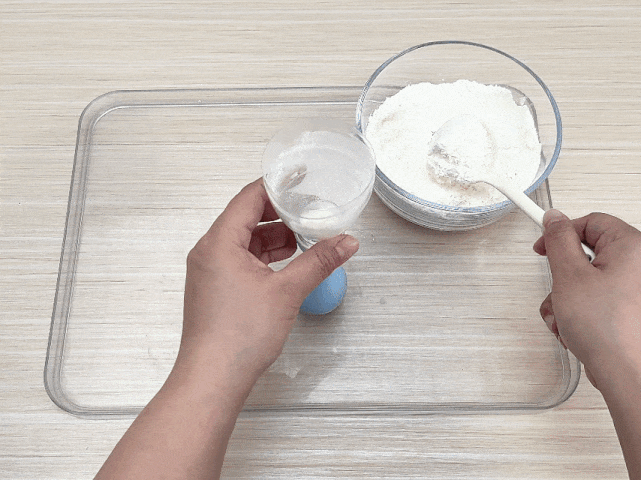 Two hands engage in a simple kitchen task on a clear plastic tray placed on a light wooden table. One hand holds a clear cup containing a white liquid, possibly water or milk, while the other hand uses a white spoon to scoop flour from a clear bowl. The scene suggests the initial steps of preparing a dough or batter.