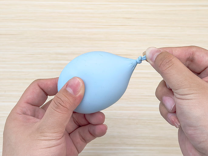 Two hands hold a small, pale blue balloon against a light wood-grain background. The balloon is tied off at the neck and appears slightly inflated, giving it a somewhat oblong shape. One hand supports the body of the balloon, while the other hand pinches the knotted end.
