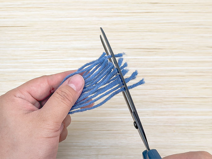 Against a light wood-grain background, two hands are shown using scissors to trim a bundle of blue yarn. One hand holds the yarn, which has been cut into numerous short strands to create a fringe-like appearance. The other hand holds a pair of blue-handled scissors, actively trimming the ends of the yarn pieces to make them even.