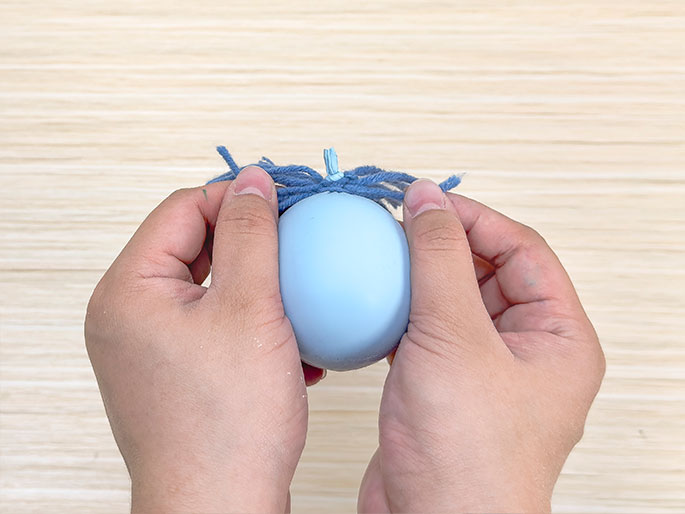 Two hands hold a pale blue balloon with a small bundle of blue yarn tied around its top against a light wood-grain background. The yarn is spread out, resembling a small tuft or tassel. The hands gently grip the sides of the balloon, which appears to be inflated to a moderate size.