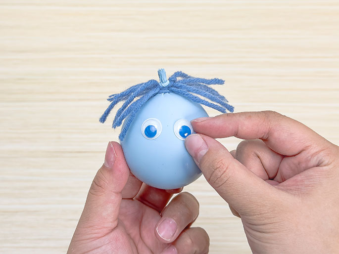 Two hands are shown in the process of creating a simple figure out of a pale blue balloon against a light wood-grain background. The balloon has a tuft of blue yarn attached to the top, resembling hair. One hand holds the balloon while the other hand applies two large, circular googly eyes with bright blue irises to the balloon's surface, giving it a face.