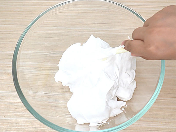 A clear glass bowl containing a thick, white, creamy substance sits on a light wood-grain surface. A hand holding a small, light-colored stick or spatula is actively stirring the mixture. This image likely depicts a step in a recipe or craft project involving mixing ingredients.