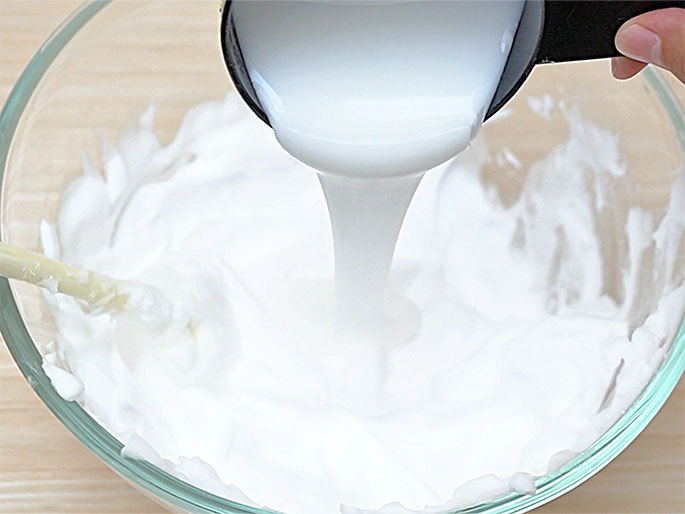 A white, viscous liquid is being poured from a black measuring cup into a clear glass bowl containing a thick, white, creamy substance. A light-colored mixing stick is partially visible in the bowl, resting on the surface of the creamy mixture. The scene is set against a light wood-grain background and suggests a step in a recipe or craft project where ingredients are being combined.