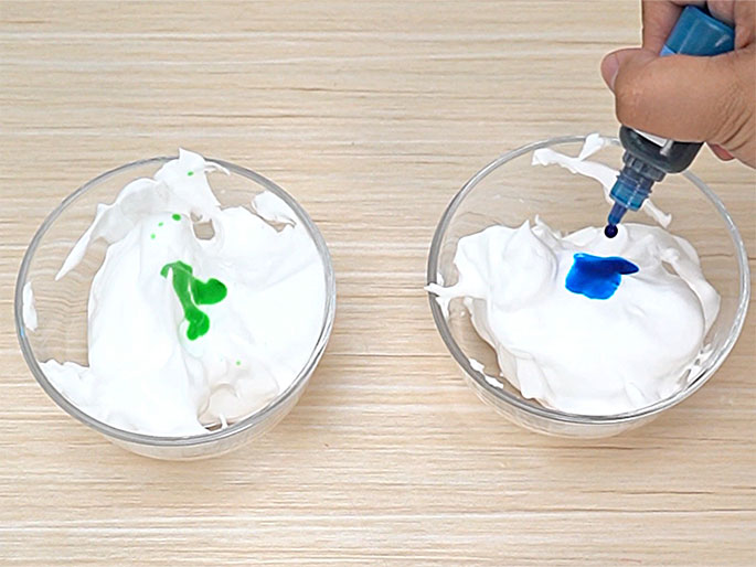 Two clear glass bowls containing a white, fluffy substance sit on a light wood-grain surface. In the left bowl, a few drops of green coloring have been added. In the right bowl, a hand is squeezing a small blue bottle, adding blue coloring to the white substance. This image likely depicts a step in a craft or cooking activity where color is being added to a mixture.