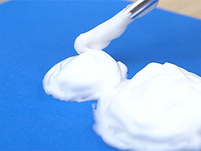 Several dollops of a thick, white, creamy substance are arranged on a bright blue surface. A metal tool, possibly tweezers or tongs, is lifting a portion of the cream, creating a swirl. The scene suggests a crafting or cooking activity.