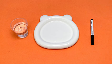 Against a bright orange background, a simple arrangement of craft supplies is presented. A small, clear plastic cup containing a quantity of clear liquid sits to the left. A white ceramic dish, shaped like a bear's face, is positioned in the centre. To the right of the dish is a black permanent marker. The scene is clean and well-lit, suggesting preparation for a craft project.