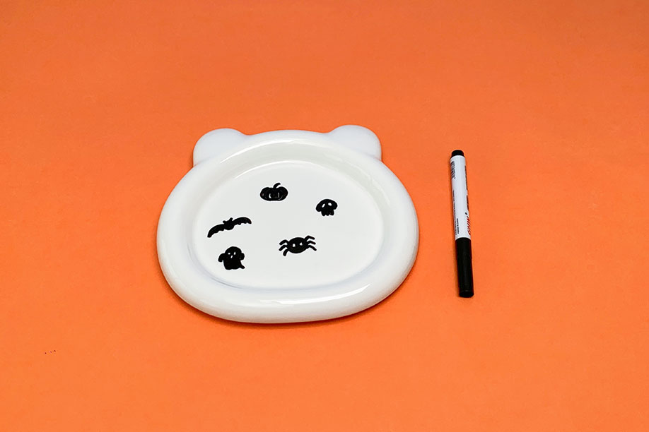 A white ceramic dish, shaped like a bear's face, rests on a bright orange surface. Several small, black Halloween-themed images—a ghost, bat, spider, pumpkin, and skull—have been drawn onto its surface using a black marker pen, which is also shown in the image. The simple, childlike drawings are neatly executed, creating a charming and festive effect. The overall impression is one of a simple, fun craft project.