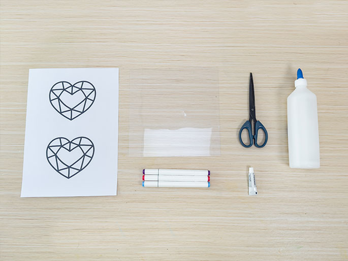Arranged on a light wood-grain surface are various craft supplies. These include a piece of paper with two printed outlines of faceted, heart-shaped gems, a clear plastic sheet with a small amount of white material inside (possibly glue or glitter), a pair of blue-handled scissors, a white bottle of glue, three markers with purple, red, and light blue caps, and a small tube of adhesive.