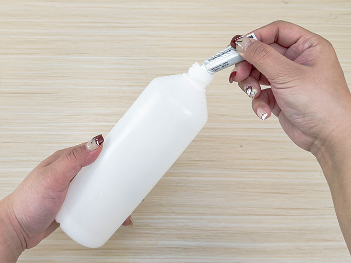 Against a light wood-grain background, two hands are depicted adding adhesive to a larger container. One hand holds a white plastic bottle at an angle, while the other hand squeezes a small, silver tube of glue or adhesive into the bottle's opening. The fingernails on the hand holding the tube have decorative nail art.