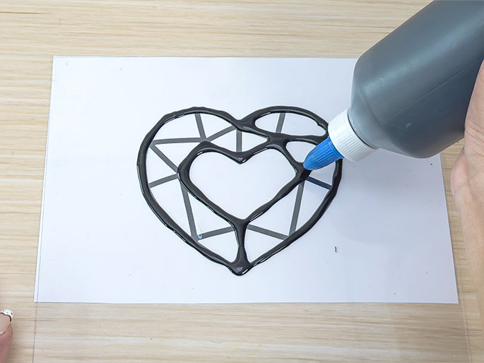 A hand holding a squeeze bottle with a blue tip is applying thick, black glue or paint to a printed design on a piece of white paper placed on a clear plastic sheet. The design is a heart shape with geometric facets resembling a gem. The glue or paint is being applied along the lines of the design, creating a raised, outlined effect. The background is a light wood-grain surface.