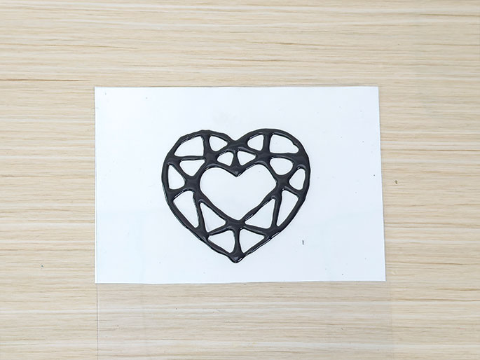 A raised, black outline of a faceted heart gem, created with glue or paint, sits on a white piece of paper atop a clear plastic sheet. The black material follows the lines of a pre-printed design, giving it a three-dimensional appearance. The background is a light wood-grain surface.