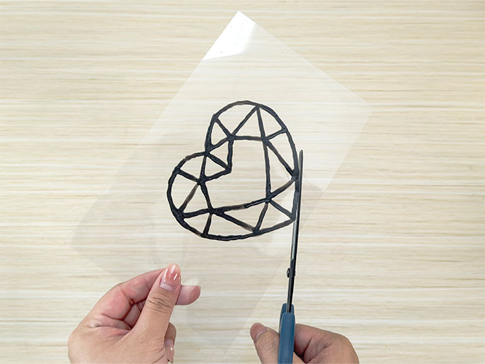 Two hands are shown cutting a dried, raised outline of a faceted heart gem from a clear plastic sheet. The outline, made of black glue or paint, is being carefully trimmed with blue-handled scissors. The background is a light wood-grain surface.