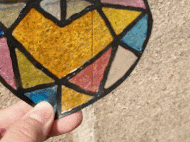 A close-up of a hand holding a heart-shaped faux stained glass piece. The heart is made up of different geometric shapes in bright colors such as yellow, pink, blue, and beige, all outlined in black. The camera focuses on the details of the colors and textures inside the heart as the hand slightly adjusts its position. The background is a textured, light gray surface, most likely a wall or floor.