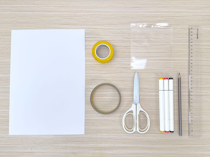 A sheet of white paper, rolls of yellow and clear tape, a clear plastic bag, scissors, white markers, a pencil, and a ruler are arranged on a light wood-grain surface.