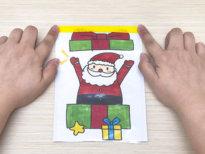 Two hands hold a clear plastic bag containing a drawing of Santa Claus popping out of a present, with a strip of yellow tape across the top of the bag, against a light wood-grain background.