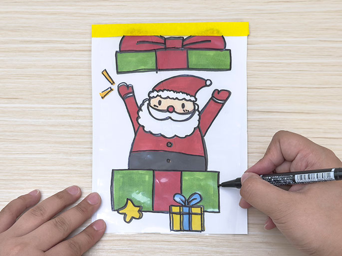 A hand colours in a drawing of a present beneath a cartoon Santa Claus, enclosed in a clear plastic bag with yellow tape at the top, on a light wood-grain surface.