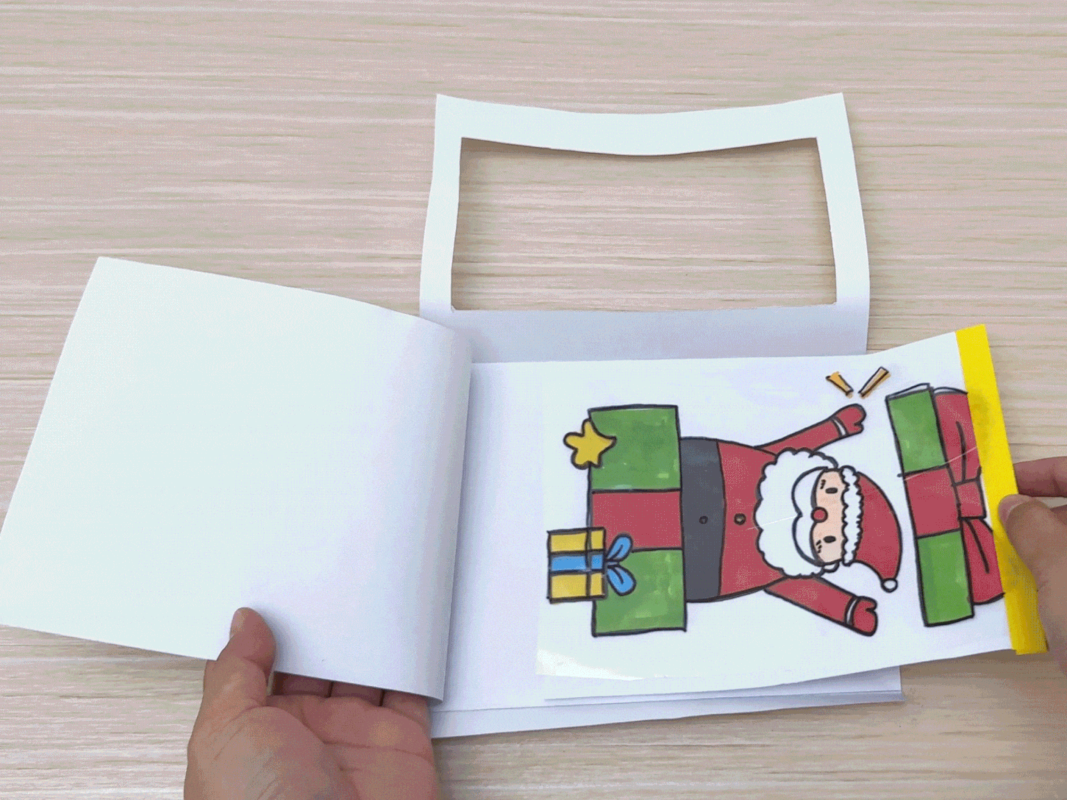 Two hands arrange a piece of white paper with a rectangular window cut out over a drawing of Santa Claus, which is sealed in a plastic bag with yellow tape, on a light wood-grain surface.