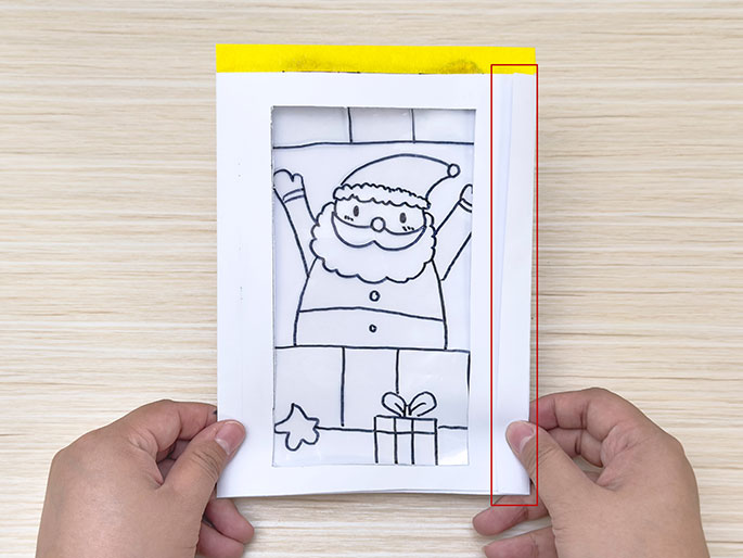 Two hands hold a white card with a yellow top edge, featuring a cut-out window revealing a black and white drawing of Santa Claus, a present, and a star, against a plain, light wood-grain background.