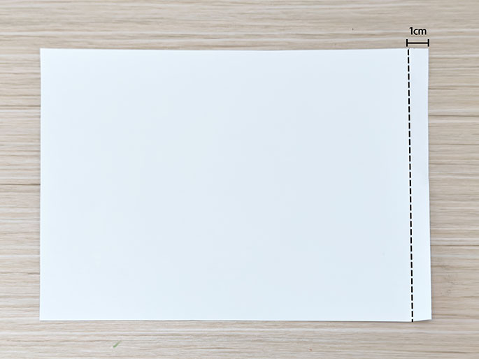A sheet of white paper on a light wood-grain surface has a dashed line drawn one centimeter from the right-hand edge.
