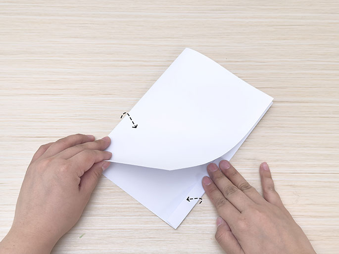 Two hands fold a sheet of white paper along a dashed line, one centimeter from the right-hand edge, on a light wood-grain surface.