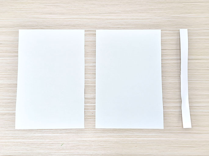 Two identical rectangular pieces of white paper and a long, thin strip of white paper are arranged on a light wood-grain surface.