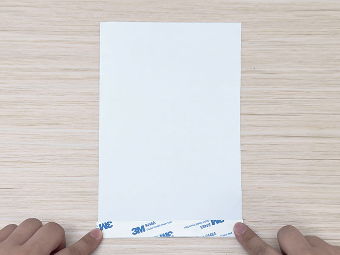 Two hands hold a piece of white paper on a light wood-grain surface, with a strip of double-sided tape adhered along the bottom edge.