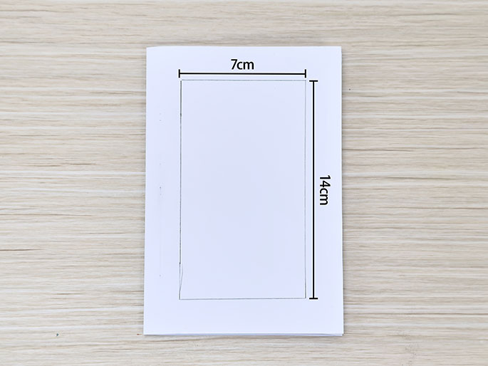 A rectangular piece of white paper on a light wood-grain surface has a smaller rectangle drawn in the center, measuring 7cm wide and 14cm high.