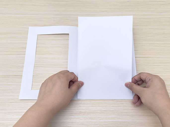 Two hands hold a piece of white paper with a rectangular cut-out window next to another piece of white paper on a light wood-grain surface.