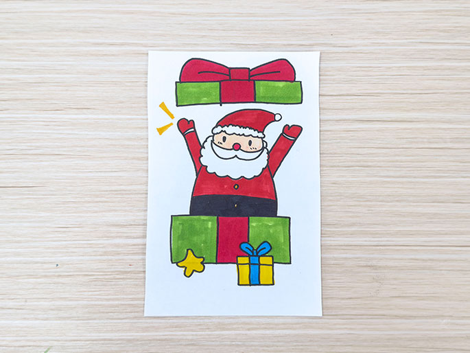 A drawing on white paper, placed on a light wood-grain surface, depicts a cartoon Santa Claus popping out of a green and red present, with another present and a star drawn below and the top of a present drawn above.
