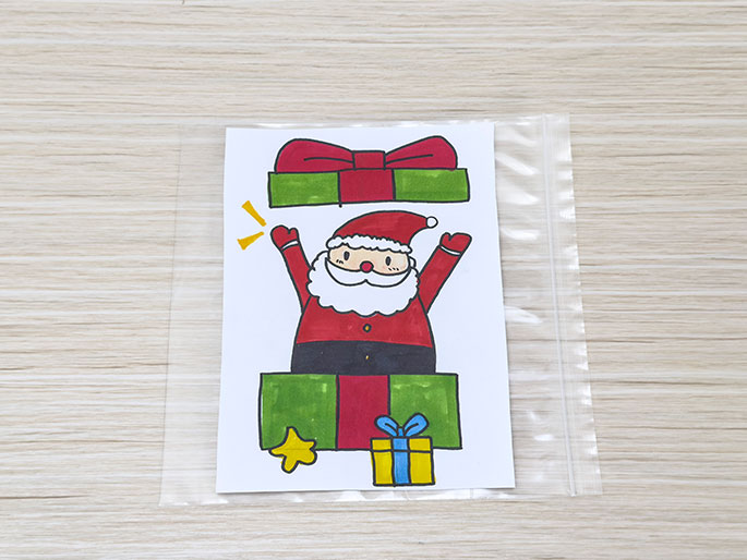 A drawing on white paper, depicting a cartoon Santa Claus popping out of a present, is sealed inside a clear plastic bag on a light wood-grain surface.