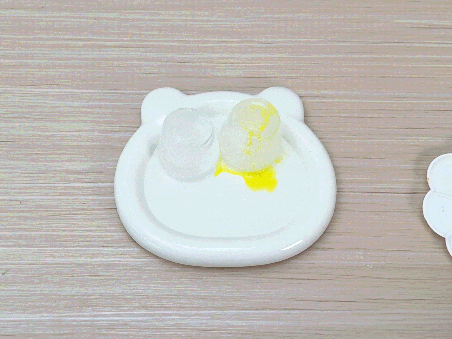Two ice cubes sit on a small, white, bear-shaped dish on a wooden table. One ice cube is clear, while the other has thin, orange lines on it, and a small puddle of yellow liquid has formed around its base. A hand holding a paintbrush with green paint then paints the clear ice cube.