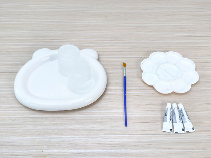 Arranged on a light wood-grain surface are several art supplies. A white, bear-shaped dish holds two small, clear containers, likely for water or mixing mediums. Next to it lies a small blue paintbrush with a tan bristle tip. A white, flower-shaped paint palette with small wells for paint sits to the right of the brush. Three small, unopened tubes of paint or glue, featuring gray and white packaging, are grouped together at the far right. This image depicts a set of materials ready for a painting or craft project.