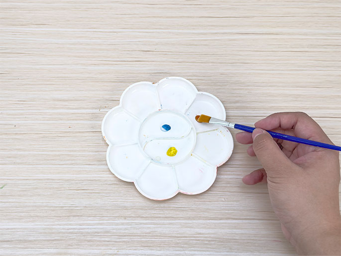 A white, flower-shaped paint palette rests on a light wood-grain table. Small dabs of blue and yellow paint are in two of the palette's wells. A hand holding a small blue paintbrush with a tan bristle tip is dipping the brush into the yellow paint, preparing to mix or apply the color. This close-up image suggests the beginning stages of a painting or craft activity.