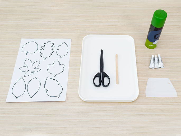 An overhead flatlay image displays craft supplies arranged on a light woodgrain table. These supplies include a sheet of paper with various leaf outlines printed on it, a pair of black scissors, a wooden craft stick, a small green and dark blue can labeled FOA three small tubes of adhesive, and a small white spatula or scraper. A white rectangular tray holds the scissors and craft stick. The arrangement suggests preparation for a leaf-themed craft project.