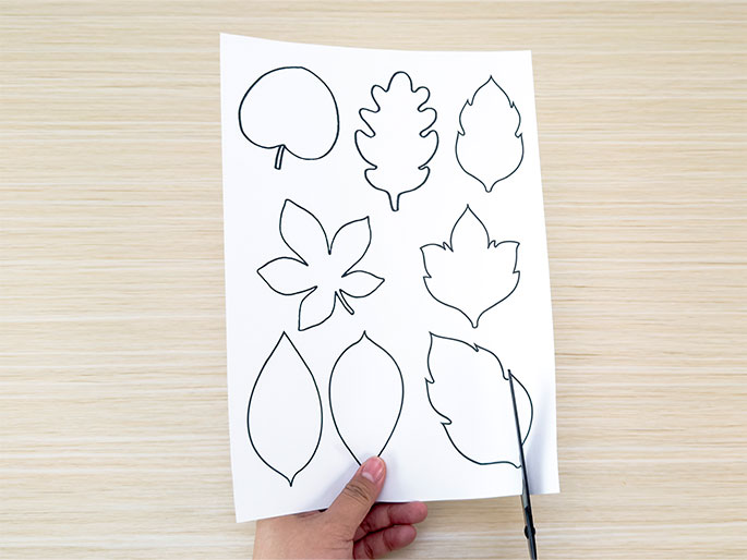 A hand holds a piece of white paper with outlines of various leaf shapes printed on it against a light woodgrain background. A pair of scissors is cutting out one of the leaf shapes from the bottom right corner of the paper. The image suggests the beginning of a craft project involving paper leaf cutouts.