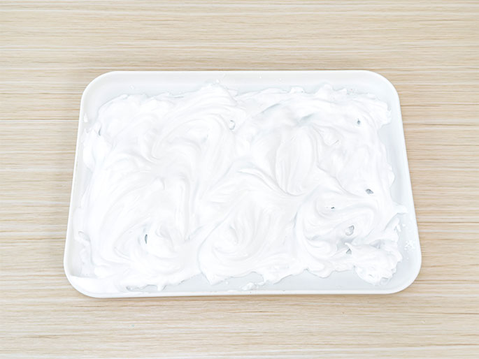 A white rectangular tray on a light woodgrain table holds a swirled mass of white foam or cream. The texture appears light and fluffy. The image suggests a craft or sensory activity, possibly involving shaving cream, whipped cream, or a similar substance.