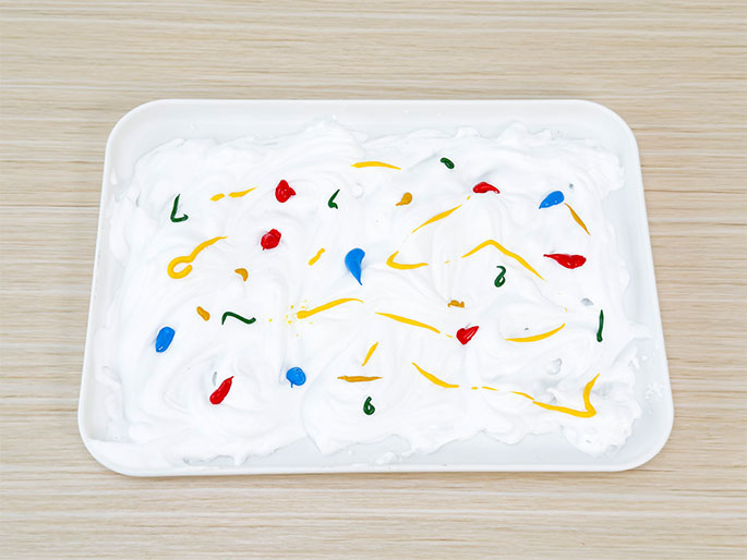 A white rectangular tray sits on a light woodgrain table. Inside the tray, a layer of white foam or cream is swirled, and drops of yellow, red, blue, and green paint have been added to the surface. The paint appears to be slightly mixed in some areas, creating streaks of color. This setup suggests a sensory or art activity, possibly marbling or creating patterns with paint and foam.