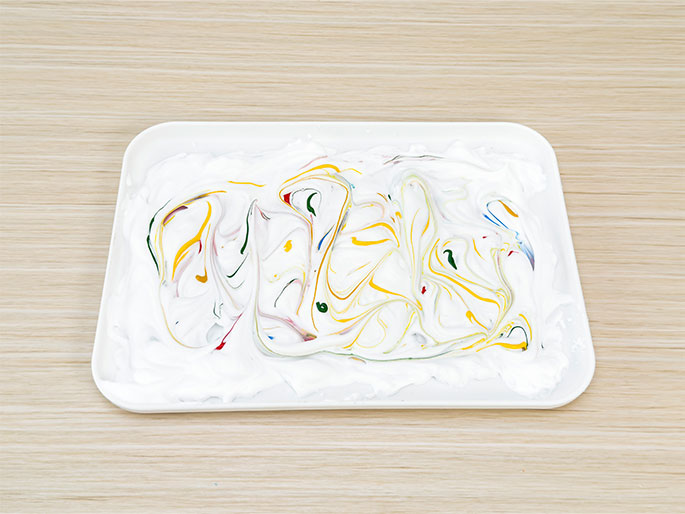 On a light woodgrain table, a white rectangular tray contains a bed of white foam or cream. Swirls of yellow, red, blue, and green paint have been marbled throughout the foam, creating a vibrant and abstract pattern. The image suggests a marbling art activity.