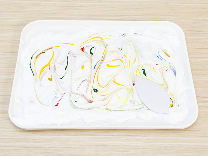 A white rectangular tray rests on a light woodgrain table. The tray contains white foam or cream, marbled with swirls of yellow, red, blue, and green paint. A small white spatula or scraper is partially submerged in the foam on the right side, suggesting it was used to create the marbled effect. The scene depicts a marbling art activity in progress.