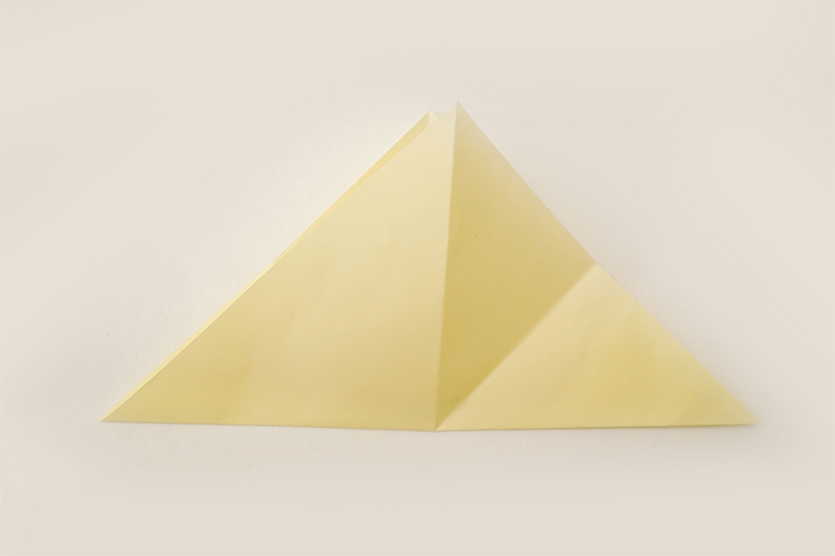 A pale yellow square of paper is folded in half diagonally, creating a crisp isosceles triangle. The fold line runs precisely down the centre, dividing the triangle into two symmetrical halves. The paper lies flat on a plain white background, the even lighting highlighting the clean fold and the uniform colour of the paper. The image is simple and straightforward, clearly showing a single, precise origami fold.