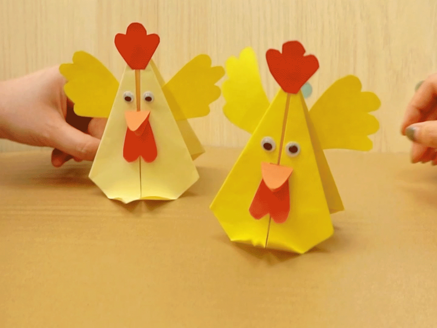 Two cheerful, origami chickens stand on a light brown surface. Each chicken is primarily yellow, with bright red combs and wattles, and small, black googly eyes. They have simple, yellow paper wings. A hand is visible near one of the chickens, perhaps adjusting its position. The chickens are charmingly simplistic in design, suggesting a completed craft project. The overall scene is bright, clean, and conveys a sense of accomplishment.