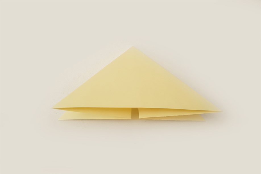 A pale yellow piece of paper, initially folded into a triangle, has been further manipulated. The top point of the triangle is now folded downwards, creating a smaller, inverted triangle at the apex and forming distinct layers of folded paper. The piece lies flat on a plain white background, the even lighting clearly showing the precise folds and the uniform colour. The image depicts a specific stage in a paper-folding process, likely origami.