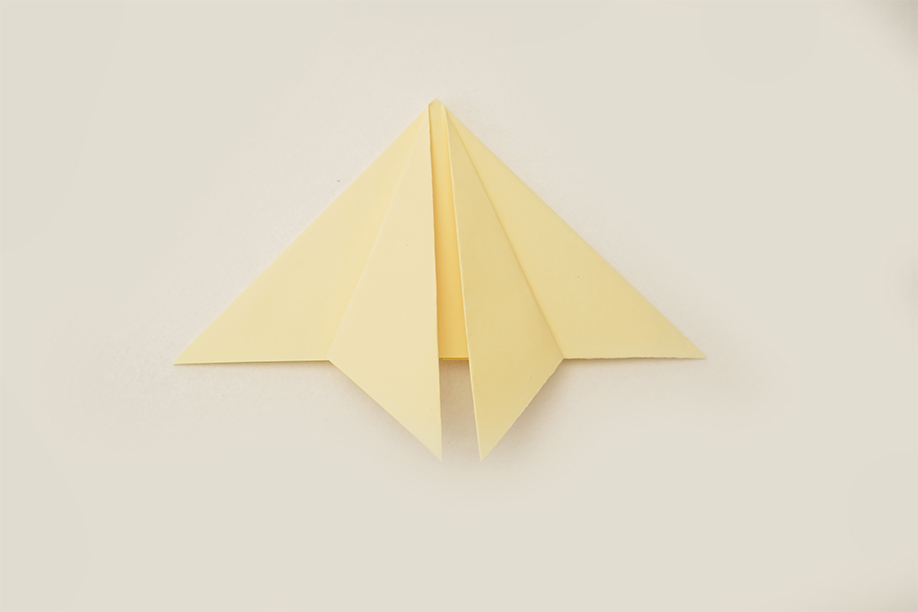 A pale yellow square of paper, following previous folds, has been further manipulated into a more complex shape. The paper now resembles a pointed, symmetrical kite or a simplified bird form, with sharp angles and clean creases. The piece lies flat on a plain white background, the even lighting clearly showing the precise folds and the uniform colour. The image depicts a clear step in a paper-folding process, likely origami, showing a more advanced stage than previous folds.